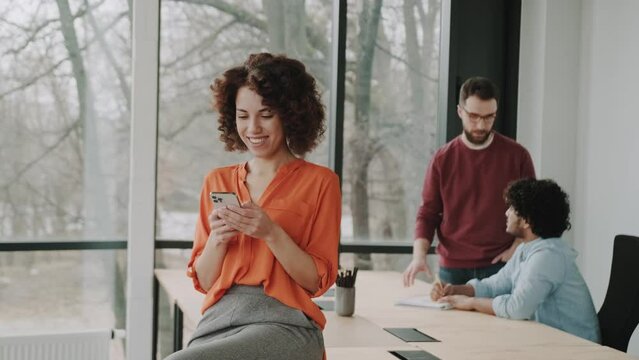 Happy millennial African American woman in the office holding smartphone using ecommerce apps, playing mobile games, chatting in messengers or social media networks, surfing internet or texting