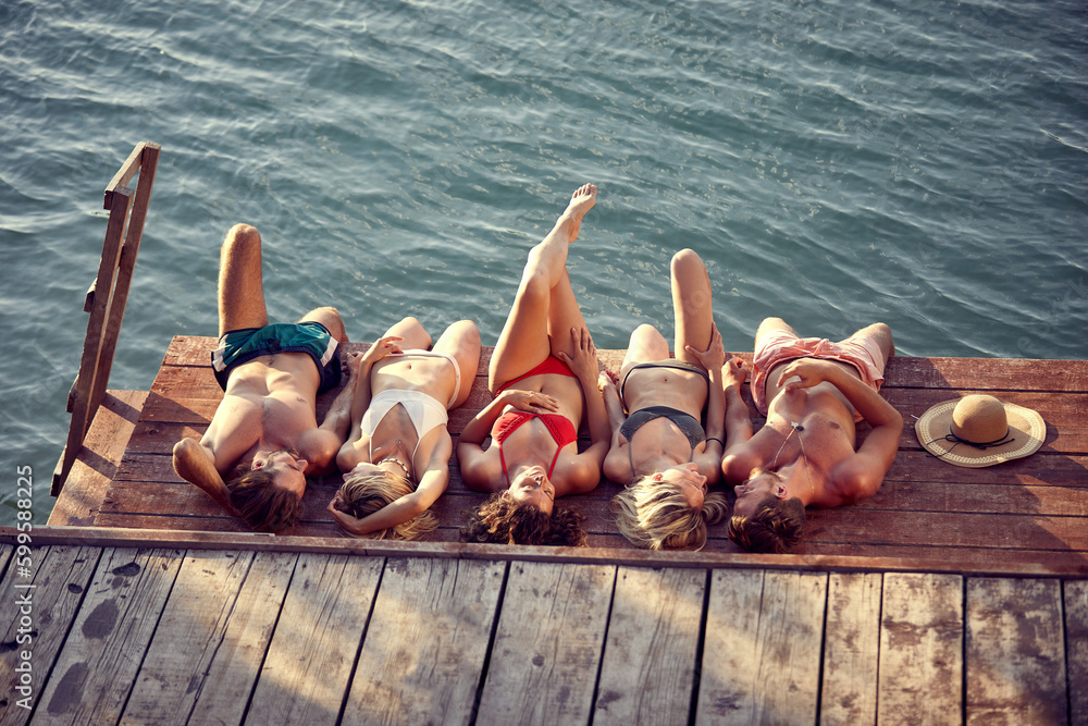 Wall mural Group of young friends laying on a wooden jetty by water, sunbathing. Young men and women on summer vacation. Holiday, fun, togetherness, lifestyle concept.