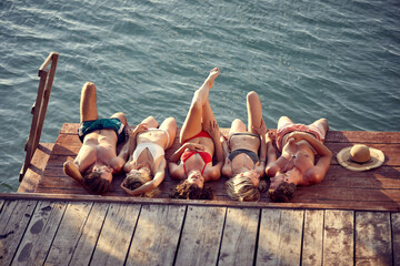 Group of young friends laying on a wooden jetty by water, sunbathing. Young men and women on summer vacation. Holiday, fun, togetherness, lifestyle concept.