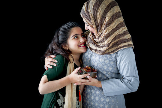 Beautiful Asian Muslim woman with daughter girl wear hijab scarf and traditional costume, hug each other, hold date fruits together, sweet food eating in Ramadan of Islam on dark black background.