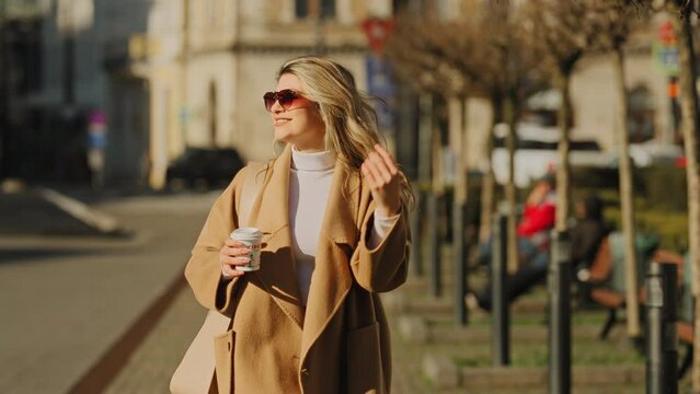 Slow motion of a young beautiful blonde woman walking with confidence and smiling, holding a takeaway coffee cup, on a sunny morning