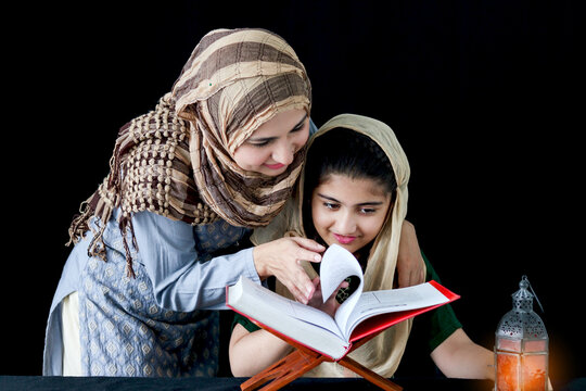 Adorable Pakistani girl with beautiful eyes wear hijab, learning Quran book with mother, Muslim family, daughter and mom read holy Koran book of Islam with brighten lantern on dark black background.