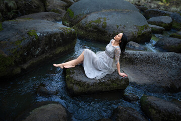 A woman sits on a rock in a river with moss on it