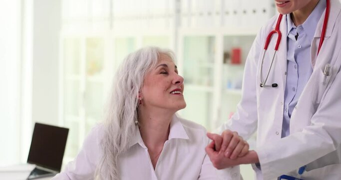 Elderly woman visits doctor and receives optimistic news after medical examination. Therapist holding hand of old patient and giving hope