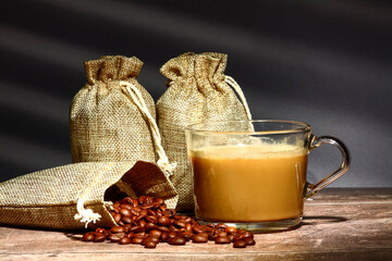 Sacks of raw roasted coffee beans and a glass cup of coffee with a reflection of a window blind in...
