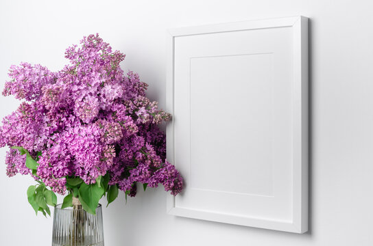 Blank vertical picture frame mockup with fresh lilac flowers bouquet in vase over white wall