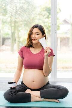 Happy beautiful young pregnant Asian woman in fitness clothes sitting cross legs on a yoga mat in her living room holding anti-stretch mark cream and gel in her hands
