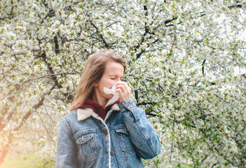 Woman sneezing into a handkerchief near a tree full of blossoms. She is suffering from seasonal...