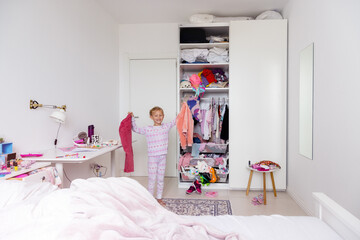 Little girl in pink pink pajamas hangs things in the closet, mess in the room