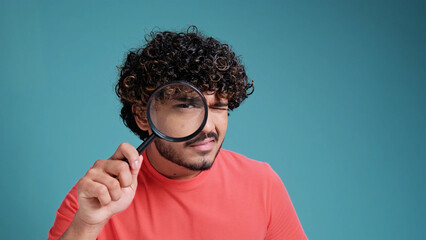 funny young latin spanish guy with a magnifying glass in his hands looks out or looks for something, dressed in casual, on a blue studio background