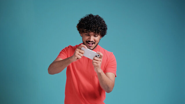 Excited happy young latin or arabian man gamer player using smartphone, playing game in mobile videogame app or watching digital video stream holding cell phone isolated on blue studio background.