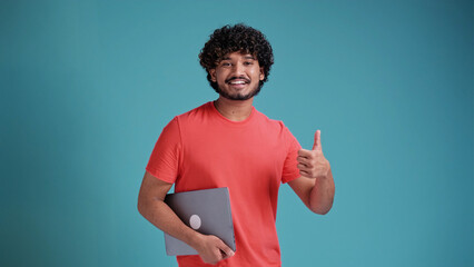 young handsome arabic or latin man showing thumbs up in a coral t-shirt on a blue background with a laptop in his hands looks at the camera and smiles