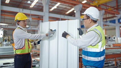 Two professional Asian man workers in safety uniform holding and checking quality of metal sheet roof at manufacturing metal plant