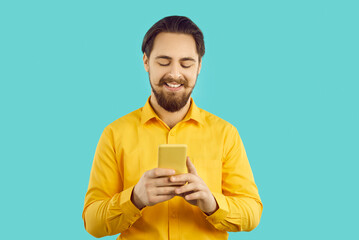Smiling young Caucasian man isolated on blue studio background text message online on mobile phone. Happy guy browse wireless internet on smartphone. Technology concept.