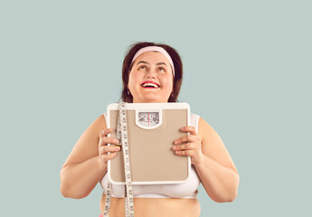 Happy, fat, overweight, chubby young woman in a sports bra standing on a light background, holding...