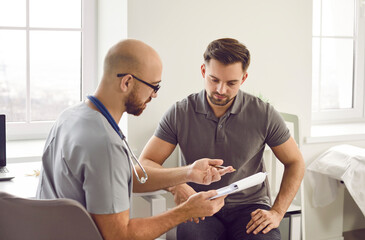 Doctor talks to his patient after a medical examination at the clinic. Young male physician shows a clipboard with the results of some medical tests they did and explains what this all means - obrazy, fototapety, plakaty