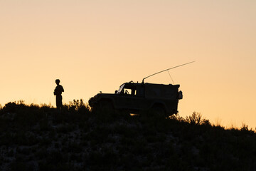 Naklejka na ściany i meble Un soldado centinela con su vehículo en el campo al atardecer.