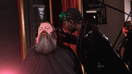 An African-American woman in a black raincoat shaves a man's beard with a straight razor in a barber shop