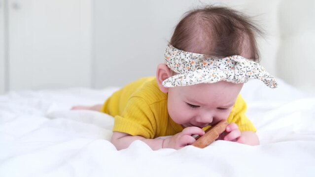 healthy asian baby girl in yellow bodysuit plays with wooden toy teether for teeth on white bedding