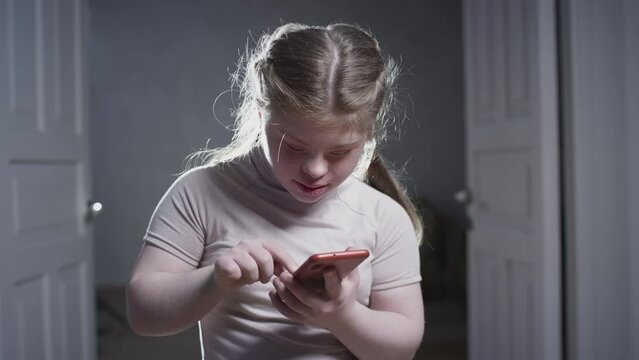 A girl with down syndrome is holding a smartphone. Writes a message, looks at photos, uses a social media app. Disabled person at home. Life with a disability.