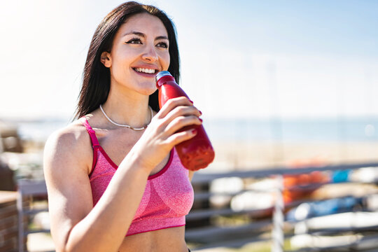 Young sporty woman wearing sportswear drinking water and relaxing after gym- Happy fitness girl holding bottle outdoors at jetty beach enjoying summer workout-Healthy Life style and Training Concept 