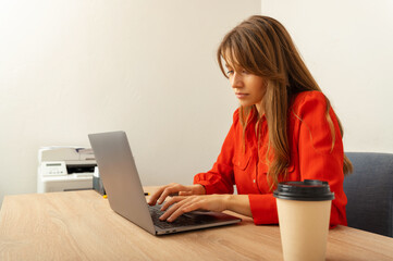 Young concentrated woman wearing red shirt is sitting in the office working hard on her laptop.