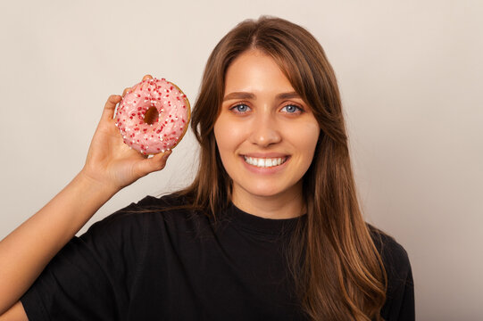 Close up portrait of a blonde woman holding a pink donut near her face over grey background.