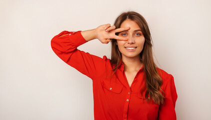 Handsome young cheerful blonde woman is holding v gesture over her eye over grey background.