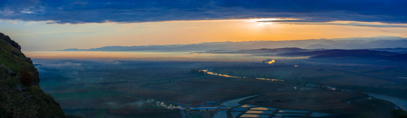 Aerial view of the valley in early morning mist, beautiful in the highlands. Low clouds and fog cover the sleeping meadow. Alpine mountain valley mists landscape at dawn. Serene moment in rural area