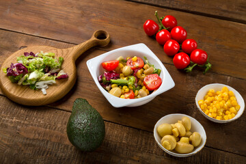 Vegetable salad with champignon lettuce and cherry tomato with avocado olive oil in a white plate on a wooden table next to all the ingredients.
