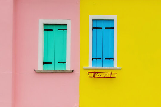 The pink facade of the house and the window with green shutters. There is also a yellow facade of the house and a window with blue shutters. Colorful houses on the island of Burano, Venice, Italy. 