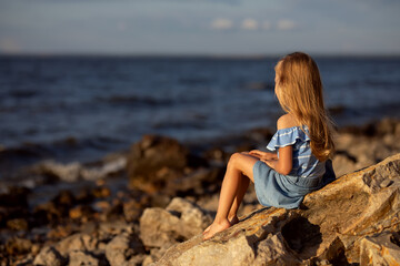 Beautiful girl with long blond hair sits on a stone on the banks of a river, lake, sea.