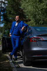 Fototapeta na wymiar Caucasian bearded man in a blue suit gets out of a black electro car in the countryside in summer.