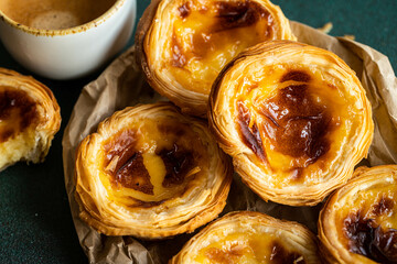 Pile of fresh traditional Portuguese pastel de nata on paper bag and cup of espresso on green surface.