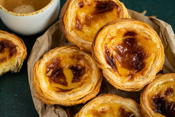 Pile of fresh traditional Portuguese pastel de nata on paper bag and cup of espresso on green surface.
