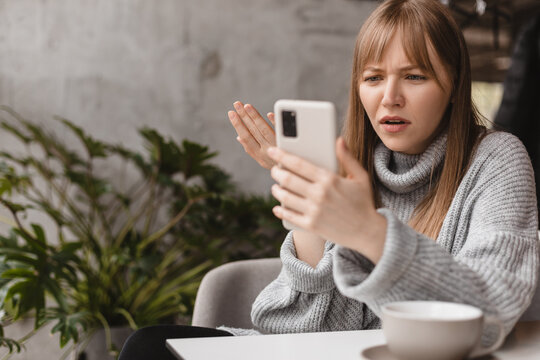 Confused puzzled blonde bang woman in casual clothes, sits in cafe, holds a smartphone in her hand, looks questioningly at the mobile phone, spreading her arm around. Girl getting surprising bad news.