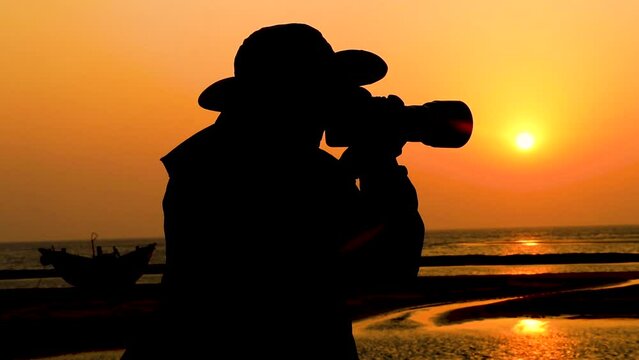 Silhouette of the photographer standing on the camera's shoulder in front of the ocean at sunset. Silhouette and back views of a travel photographer.