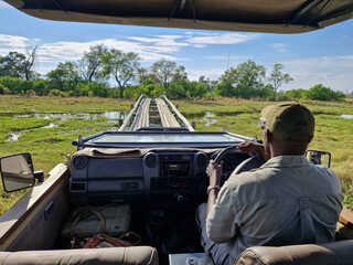 Ein Guide mit Mütze am Steuer eines Safari – Jeep fährt über eine wacklige Holzbrücke über einen Fluss im Okavango Delta von Botswana, Afrika	