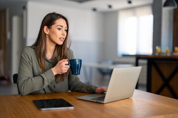 Young beautiful businesswoman using laptop computer and holding cup of coffee. Photo of female working on laptop device or reading news while drinking coffee in home office.