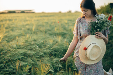 Beautiful woman with wildflowers and straw hat walking in barley field in sunset light. Stylish...