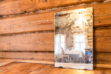 Obraz na płótnie Canvas windows, table and chairs of village house are reflected in old mirror standing on floor of log cabin (focus on the reflection in the mirror)