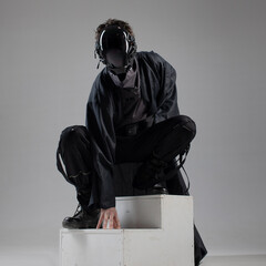 Urbantech outfit cyber style, a young man in stylish black clothes and a mirror mask on his whole face, sitting on the stairs. Studio photo on a light background - 599706299