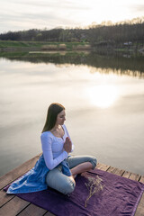 Fototapeta premium Woman relaxingly practicing meditation in nature by the lake. Nature background. Spiritual and emotional concept.