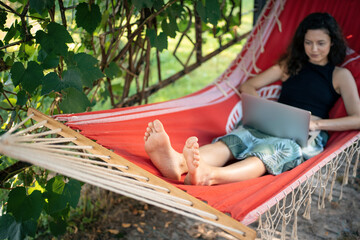 focused young female typing on laptop while lying in red hammock outside