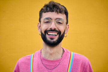 Close up smiling cheerful young Caucasian man holding his rainbow lgbt suspenders. Gay people posing for photo yellow background. Cheerful gay bearded person happy studio photo isolated. Millennials. - obrazy, fototapety, plakaty