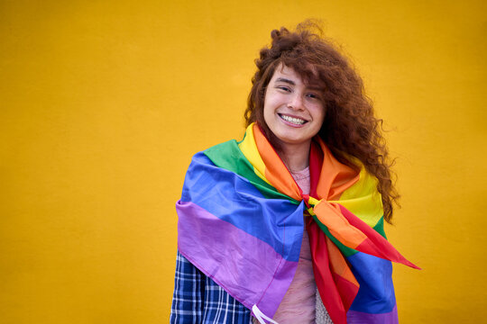 Portrait of cheerful non-binary person with red hair with rainbow flag smiling looking at camera on yellow background. Concept of LGBT community, diversity, transgender and non-binary people. 