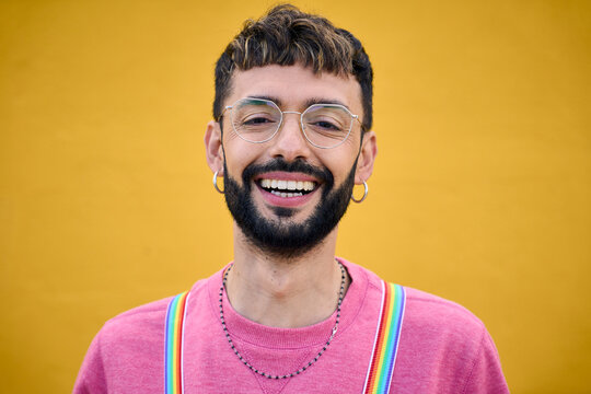 Close up smiling cheerful young Caucasian man holding his rainbow lgbt suspenders. Gay people posing for photo yellow background. Cheerful gay bearded person happy studio photo isolated. Millennials.