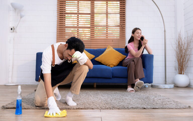 Young Asian man cleaning the house while his wife talks on the phone on the sofa.