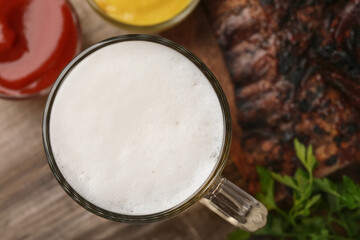 Mug with beer, delicious grilled ribs and sauces on wooden table, closeup