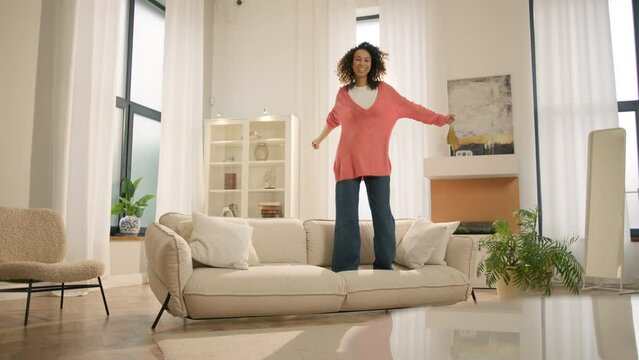 Happy young black woman jumping on sofa, feels overjoyed by celebrating move in day to own home. Mixed race woman laughing enjoying newly rented modern flat or urban apartment. Fashion furniture store